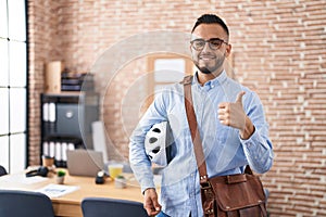 Young hispanic man working at the office holding bike helmet smiling happy and positive, thumb up doing excellent and approval