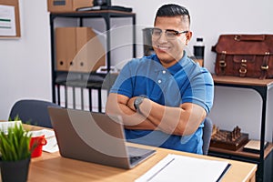 Young hispanic man working at the office with laptop happy face smiling with crossed arms looking at the camera