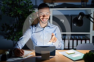 Young hispanic man working at the office at night smiling cheerful offering palm hand giving assistance and acceptance