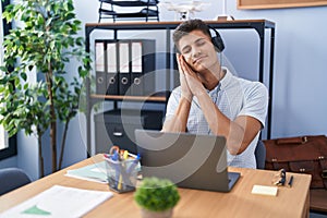 Young hispanic man working at the office wearing headphones sleeping tired dreaming and posing with hands together while smiling