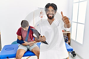 Young hispanic man working at pain recovery clinic with a man with broken arm approving doing positive gesture with hand, thumbs