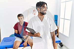 Young hispanic man working at pain recovery clinic with a man with broken arm looking away to side with smile on face, natural