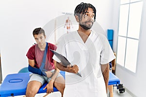 Young hispanic man working at pain recovery clinic with a man with broken arm smiling looking to the side and staring away