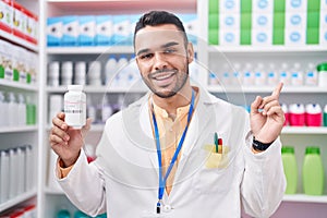 Young hispanic man working at pharmacy drugstore holding pills smiling happy pointing with hand and finger to the side