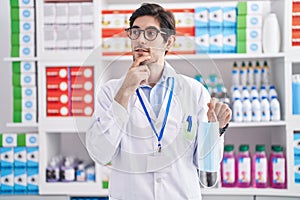 Young hispanic man working at pharmacy drugstore holding safety mask serious face thinking about question with hand on chin,