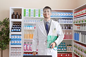 Young hispanic man working at pharmacy drugstore holding stethoscope looking positive and happy standing and smiling with a