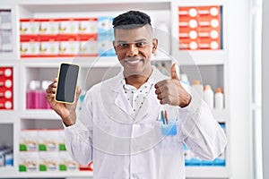 Young hispanic man working at pharmacy drugstore showing smartphone screen smiling happy and positive, thumb up doing excellent