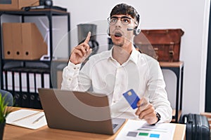 Young hispanic man working using computer laptop holding credit card amazed and surprised looking up and pointing with fingers and