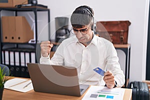 Young hispanic man working using computer laptop holding credit card angry and mad raising fist frustrated and furious while