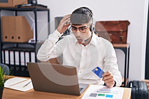 Young hispanic man working using computer laptop holding credit card crazy and scared with hands on head, afraid and surprised of