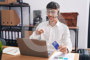 Young hispanic man working using computer laptop holding credit card pointing to you and the camera with fingers, smiling positive