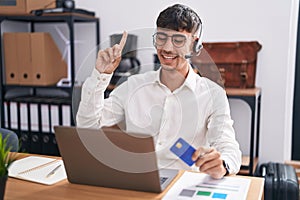 Young hispanic man working using computer laptop holding credit card smiling amazed and surprised and pointing up with fingers and