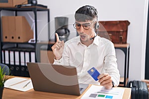 Young hispanic man working using computer laptop holding credit card surprised pointing with finger to the side, open mouth amazed