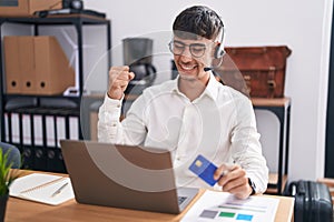 Young hispanic man working using computer laptop holding credit card very happy and excited doing winner gesture with arms raised,
