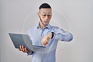 Young hispanic man working using computer laptop looking at the watch time worried, afraid of getting late