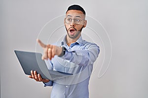 Young hispanic man working using computer laptop pointing with finger surprised ahead, open mouth amazed expression, something on