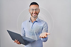 Young hispanic man working using computer laptop smiling cheerful offering palm hand giving assistance and acceptance