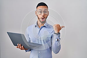 Young hispanic man working using computer laptop surprised pointing with hand finger to the side, open mouth amazed expression