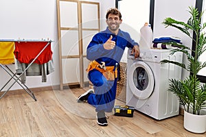 Young hispanic man working on washing machine smiling happy and positive, thumb up doing excellent and approval sign