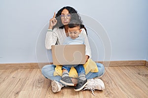 Young hispanic mother and kid using computer laptop sitting on the floor pointing up looking sad and upset, indicating direction