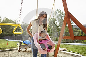 Young Hispanic mother pushing her baby on a swing at a playground in the park