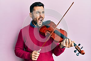 Young hispanic musician man playing violin angry and mad screaming frustrated and furious, shouting with anger