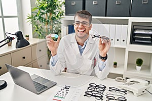 Young hispanic oculist holding glasses and contact lenses winking looking at the camera with sexy expression, cheerful and happy