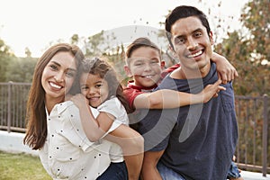 Young Hispanic parents piggyback their children in the park, smiling to camera, focus on foreground