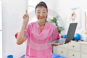 Young hispanic physiotherapist woman using computer laptop at medical clinic smiling with an idea or question pointing finger with
