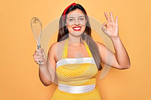 Young hispanic pin up woman wearing fashion sexy 50s style holding cooking whisk blender doing ok sign with fingers, excellent