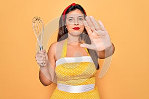 Young hispanic pin up woman wearing fashion sexy 50s style holding cooking whisk blender with open hand doing stop sign with