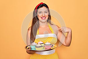 Young hispanic pin up woman wearing fashion sexy 50s style holding sweet cupcakes with surprise face pointing finger to himself