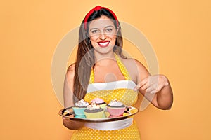 Young hispanic pin up woman wearing fashion sexy 50s style holding sweet cupcakes very happy pointing with hand and finger