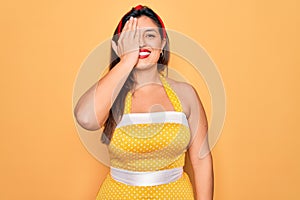 Young hispanic pin up woman wearing fashion sexy 50s style over yellow background covering one eye with hand, confident smile on