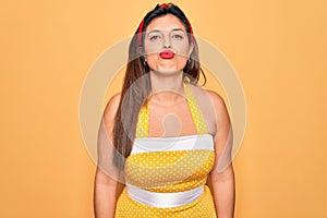 Young hispanic pin up woman wearing fashion sexy 50s style over yellow background looking at the camera blowing a kiss on air