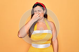 Young hispanic pin up woman wearing fashion sexy 50s style over yellow background smelling something stinky and disgusting,