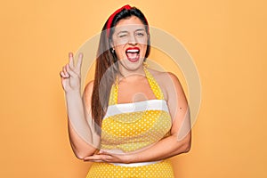 Young hispanic pin up woman wearing fashion sexy 50s style over yellow background smiling with happy face winking at the camera