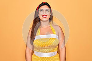 Young hispanic pin up woman wearing fashion sexy 50s style over yellow background smiling looking to the side and staring away