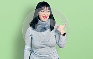 Young hispanic plus size woman wearing winter scarf smiling with happy face looking and pointing to the side with thumb up