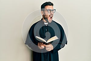 Young hispanic priest man holding bible angry and mad screaming frustrated and furious, shouting with anger