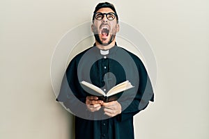 Young hispanic priest man holding bible angry and mad screaming frustrated and furious, shouting with anger looking up