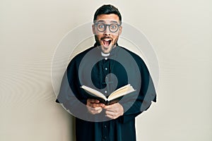 Young hispanic priest man holding bible celebrating crazy and amazed for success with open eyes screaming excited