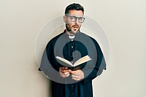 Young hispanic priest man holding bible clueless and confused expression