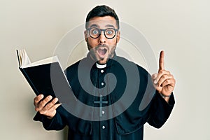 Young hispanic priest man holding bible with finger up afraid and shocked with surprise and amazed expression, fear and excited