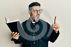 Young hispanic priest man holding bible with finger up angry and mad screaming frustrated and furious, shouting with anger