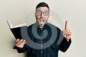 Young hispanic priest man holding bible with finger up celebrating crazy and amazed for success with open eyes screaming excited