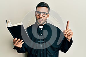 Young hispanic priest man holding bible with finger up clueless and confused expression