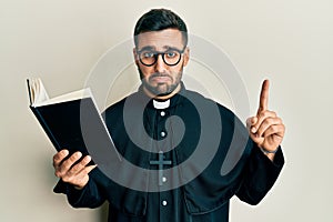 Young hispanic priest man holding bible with finger up depressed and worry for distress, crying angry and afraid