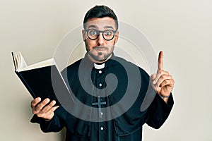 Young hispanic priest man holding bible with finger up puffing cheeks with funny face