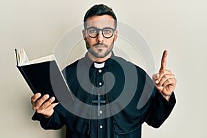 Young hispanic priest man holding bible with finger up relaxed with serious expression on face